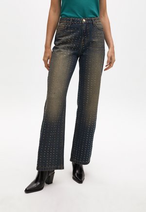 Jean en denim foncé avec une coupe droite, ornés de clous métalliques en bronze disposés en lignes verticales, et un style classique à cinq poches.