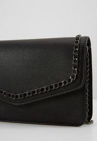 Cartera negra de piel sintética con superficie texturizada, presenta un acento de cadena a lo largo de la solapa. Forma rectangular compacta con bordes cosidos.