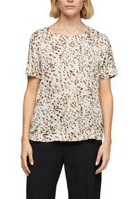 Blusa leggera a maniche corte in crema con macchie marroni sparse. Collo rotondo, vestibilità rilassata, texture liscia. Abbinata a pantaloni neri.