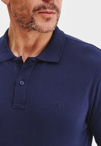 Granatowa koszulka polo wykonana z fakturowanej tkaniny, z plisą na trzy guziki i dyskretnym logo na lewej piersi.