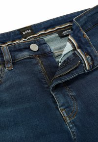 Jeans de mezclilla de color azul oscuro con la cremallera y el botón desabrochados, mostrando la tela interior y las etiquetas de la marca "BOSS" y "DELANO Slim Fit Tapered."