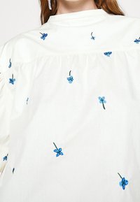 White cotton blouse featuring blue embroidered flowers, with a gathered yoke and a subtle texture. Sleeves finish with a soft cuff.