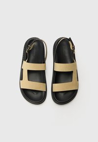 Sandales en daim tan avec deux larges sangles, un bout ouvert et une semelle en caoutchouc noir. Elles comportent une sangle réglable et un design plat.