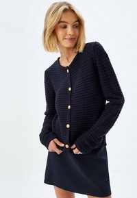 Mörkblå stickad kofta med texturerad tyg, rund halsringning och sex knappar i guldton framtill. Långa ärmar med ribbade manchetter.