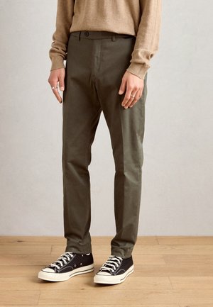 Chinos de corte slim en verde oliva, con bolsillos frontales y una textura lisa, combinados con zapatillas negras y un suéter beige.