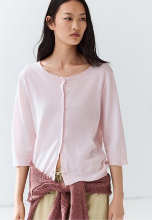 Junge Frau mit glattem dunklem Haar trägt eine hellrosa geknöpfte Strickjacke und hat einen staubrosafarbenen Pullover um die Taille gebunden.