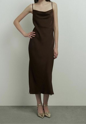 Femme portant une robe nuisette midi marron sans manches avec bretelles spaghetti et des talons dorés à bout pointu, debout contre un mur clair uni.