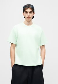 Νεαρός άνδρας που φοράει ανοιχτό πράσινο oversized t-shirt, μαύρες βερμούδες και μαύρο καπέλο γυρισμένο προς τα πίσω, στέκεται με τα χέρια στις τσέπες.