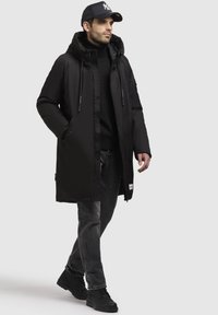 Schwarze gepolsterte Jacke mit Kapuze, ausgestattet mit einem Reißverschluss und Knopfverschluss, seitlichen Taschen und einem schwarzen Rollkragenpullover. Kombiniert mit dunklen Jeans und schwarzen Schuhen.