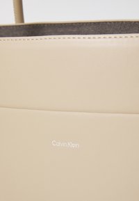 Borsa in pelle beige con texture liscia, caratterizzata da un design minimalista e un sottile logo inciso "Calvin Klein" sulla parte frontale.