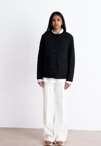 Svart vadderad jacka med knäppning, med en texturerad quiltad design och justerbara dragsnören. Bärs över en vit skjorta med krage och vida byxor.