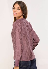 Hosszú ujjú, texturált blúz mályvaszínben, finom csíkos mintával, gallérral és gombos mandzsettákkal, sötét farmerszövethez párosítva.