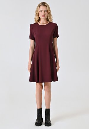 Bordeauxfarbenes Kleid mit kurzen Ärmeln, fließendem Design und subtilen Formdetails. Getragen mit schwarzen Stiefeletten. Glatte Textur.