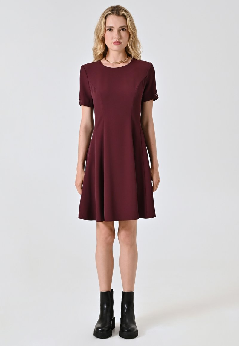 Bordeauxfarbenes Kleid mit kurzen Ärmeln, fließendem Design und subtilen Formdetails. Getragen mit schwarzen Stiefeletten. Glatte Textur.