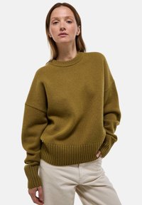 Pull en tricot vert olive avec un col rond et un ourlet en côtes. Présente des épaules tombantes et des manches longues avec des poignets en côtes.
