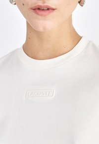 Balti medvilniniai marškiniai trumpomis rankovėmis su apvaliu iškirpimu ir iškilniu "LACOSTE" logotipu ant krūtinės. Lygus paviršius ir kasdieniškas dizainas.