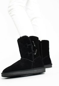Botas de tobillo de ante negro con forro de piel de oveja, que cuentan con dos cierres de toggles negros y suela de goma texturizada negra.
