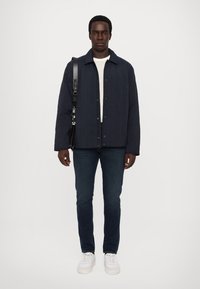 Marineblau gesteppte Jacke mit Knopfverschluss, getragen über einem weißen Pullover, kombiniert mit dunklen Jeans und weißen Sneakers. Das Modell trägt eine schwarze Tasche.