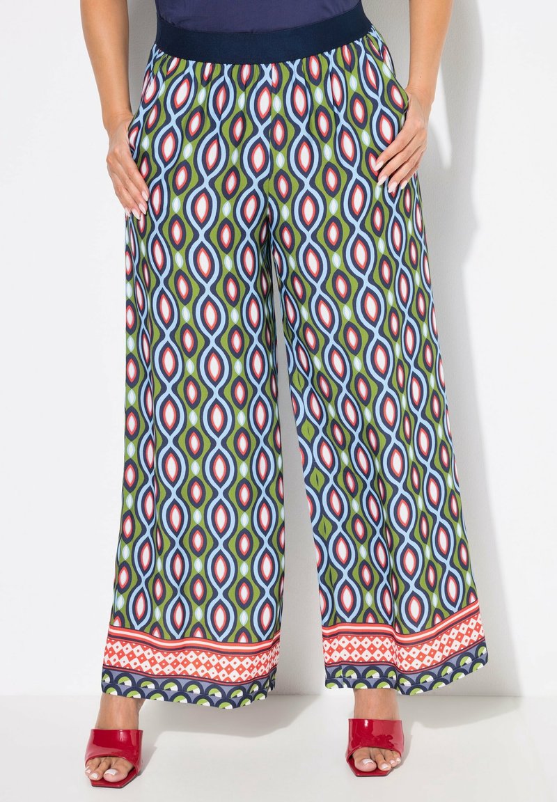 Personne portant un pantalon à jambes larges avec des motifs géométriques bleus, verts, rouges et blancs et des talons rouges ouverts à l’avant, debout les mains dans les poches.