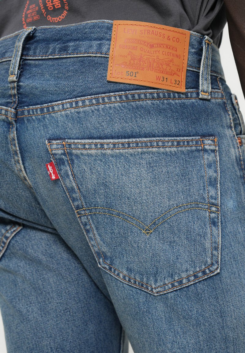 Jeans de mezclilla azul claro con un parche de cuero en la parte trasera que presenta "Levi Strauss & Co." y costuras visibles en el bolsillo, acentuados con una pequeña etiqueta roja.