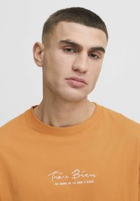Orange bomuld t-shirt med rund hals, prydet med hvid broderi, der står "Très Bien au bord de la mer d’Azur." Blød tekstur.