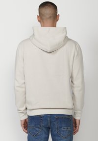 Hoodie beige chiaro con una texture morbida, dotato di tasca a marsupio e cappuccio con cordoncino, abbinato a jeans denim blu.