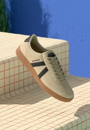 Beige sneakers med strukturerad fuskmocka, svarta detaljer och en brun gummisula. Har en logo på sidan och klassisk snörning.