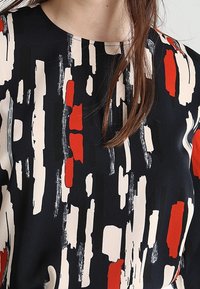 Svart topp med en djärv, abstrakt design i röda, vita och beige penseldrag; rundad hals; slät tygkvalitet.