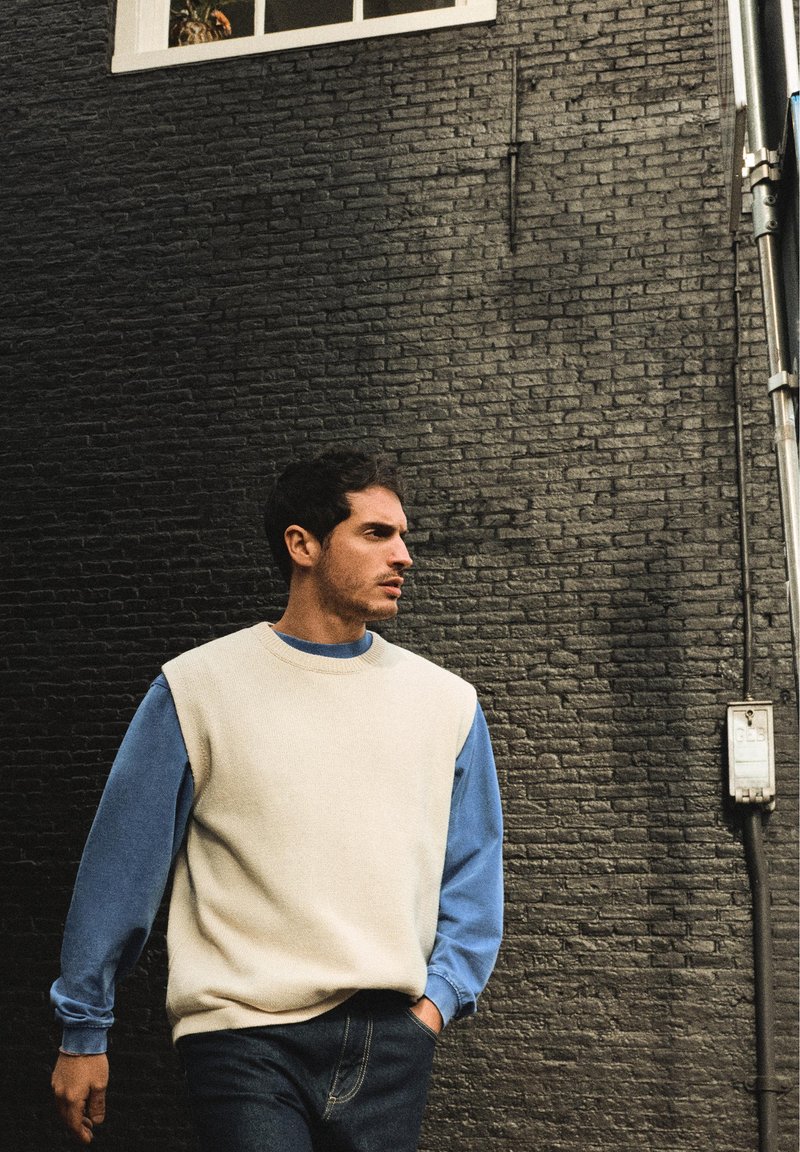 Homme aux cheveux courts et foncés portant un pull sans manches blanc par-dessus une chemise bleue à manches longues, debout contre un mur de briques sombre, les mains dans les poches.