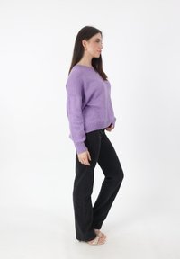 Lila gestrickter Pullover mit lockerer Passform, gerippten Bündchen und Saum, kombiniert mit hochgeschnittenen schwarzen Jeans und hellen Riemchensandalen.