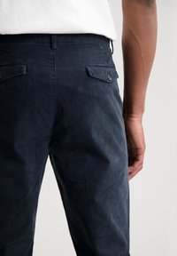 Mornice tamnoplave kord pantalone s teksturiranim završetkom, koje imaju stražnji džep i detalj gumba na pojasu. Poznate po svojoj svestranosti.