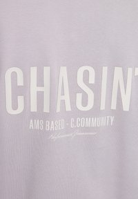 Tessuto di felpa viola chiaro con testo bianco audace: "CHASINT AMS BASED - C.COMMUNITY" con una firma in carattere più piccolo sotto.