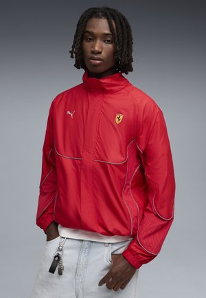 Persona con capelli ricci scuri che indossa una giacca rossa Ferrari Puma, jeans azzurri e chiavi agganciate alla tasca, su sfondo grigio.