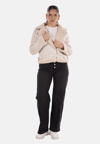 Giacca in ecopelliccia beige con chiusura a zip e colletto ampio, abbinata a pantaloni di jeans neri e sneakers bianche.