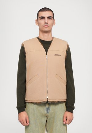 Beige ärmlös väst med dragkedja fram, två sidofickor och fransade kanter. Bärs över en mörkgrön långärmad skjorta.