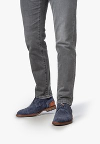 Zapatos de tela en azul marino con diseño de espiga y acentos en cuero marrón, suela plana y diseño de cordones, combinados con jeans ajustados gris.