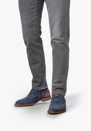 Zapatos de tela en azul marino con diseño de espiga y acentos en cuero marrón, suela plana y diseño de cordones, combinados con jeans ajustados gris.
