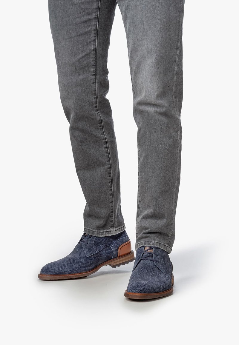 Zapatos de tela en azul marino con diseño de espiga y acentos en cuero marrón, suela plana y diseño de cordones, combinados con jeans ajustados gris.