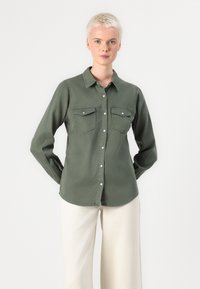 Camisa de manga longa verde-azeitona com fecho de botões, dois bolsos no peito e bainha curva. Feita de tecido leve com uma textura suave.