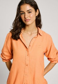 Oranges Leinen-Button-Up-Hemd mit hochgekrempelten Ärmeln, das ein dezentes Logo-Detail auf der Brust und eine lockere Passform bietet.