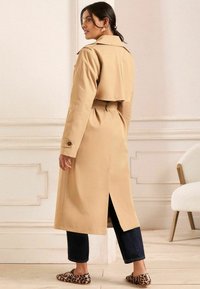 Trench coat bej deschis cu talie strânsă, epauleți pe umeri și spate despicat, asortat cu blugi închiși la culoare și loafers cu imprimeu animal.