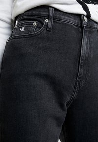 Černé džínov�é kalhoty z denimu s přiléhavým střihem, přední kapsou s logem a jemnými šitími. Textura působí hladce a odolně.