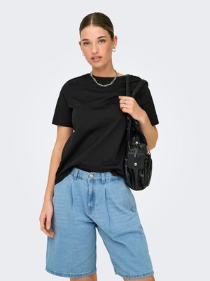 Young woman wearing black t-shirt and light blue denim shorts, carrying black handbag over shoulder against plain background.