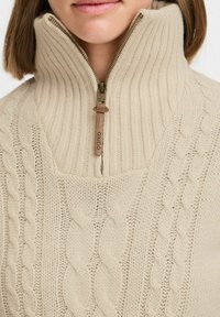 Beige Strickpullover mit hohem Kragen, der Kabelstrickmuster aufweist und einen Reißverschluss mit einem Lederzieher hat. Strukturierter, warmer Stoff.