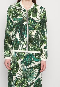 Vrouw in een bijpassende ritsjack en rok met een groot groen tropisch bladerenpatroon op een witte achtergrond.