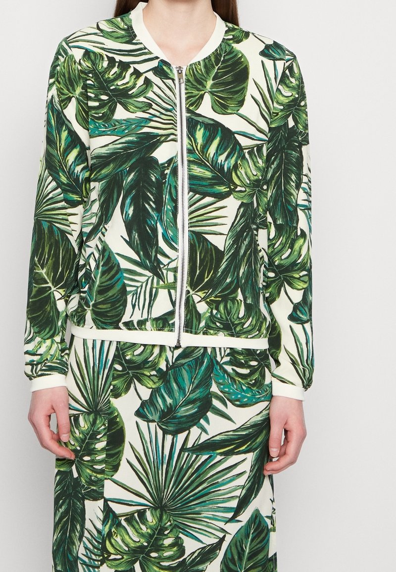 Vrouw in een bijpassende ritsjack en rok met een groot groen tropisch bladerenpatroon op een witte achtergrond.