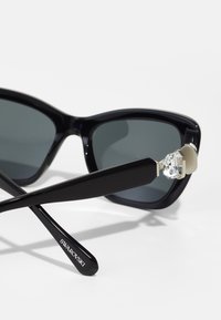 Occhiali da sole neri con design a occhio di gatto, lenti grigie e cerniera con cristalli. Le aste presentano il logo "Swarovski" in testo bianco.