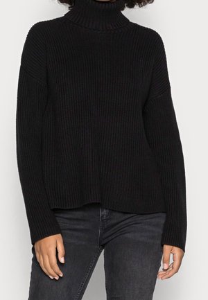 Persona vistiendo un suéter negro de cuello alto acanalado y jeans gris oscuro, mostrada desde los hombros hasta las caderas sobre un fondo liso.