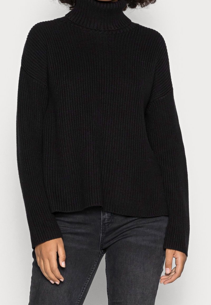 Personne portant un pull à col roulé côtelé noir et un jean gris foncé, visible des épaules aux hanches sur un fond uni.