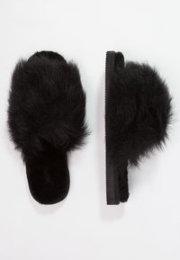 Mustat karvahuopikengät, joissa on pörröinen yläosa ja tasapohja. Materiaali on pehmeä ja teksturoitu, ja siinä on yksinkertainen, minimalistinen muotoilu.